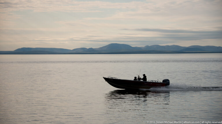 Two people travel in an open motorboat across calm water, with a low mountain range on the horizon.