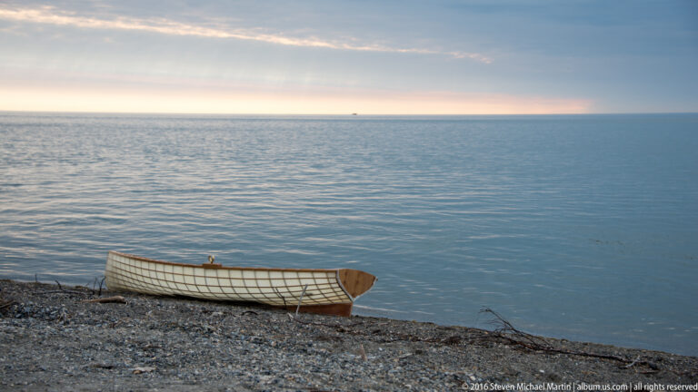 A long open boat with a pale covered frame rests on a pebbled beach beside calm water.