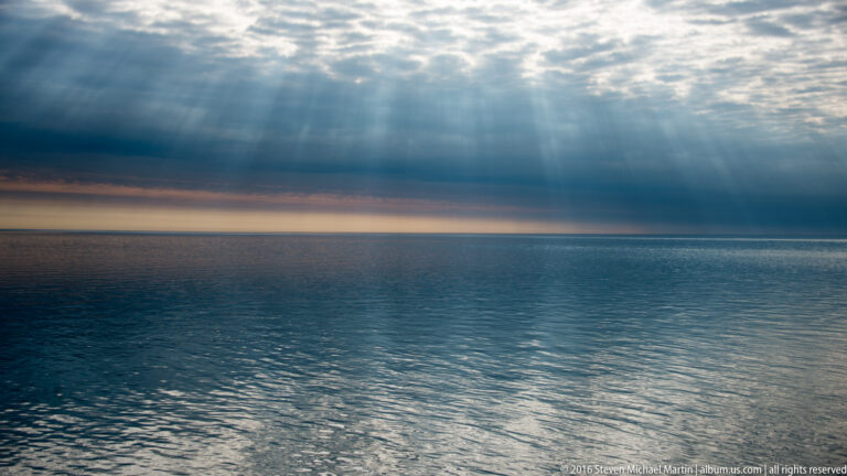 Sunbeams break through dark clouds over open water, above a thin warm band along the level horizon.