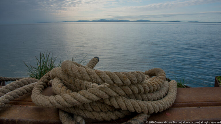 A heavy coiled rope rests on a metal edge above calm water and a low distant shoreline.