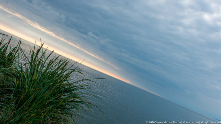 Dense grass fills the left foreground as the sea and glowing horizon tilt diagonally beneath clouds.