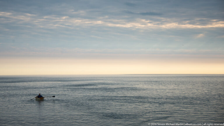 A lone person paddles a small craft across open water beneath a pastel sky and level horizon.