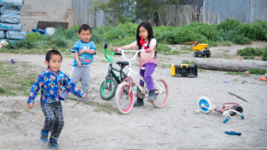 Three children gather among bicycles and toys on a sandy gravel yard bordered by shrubs and a log.