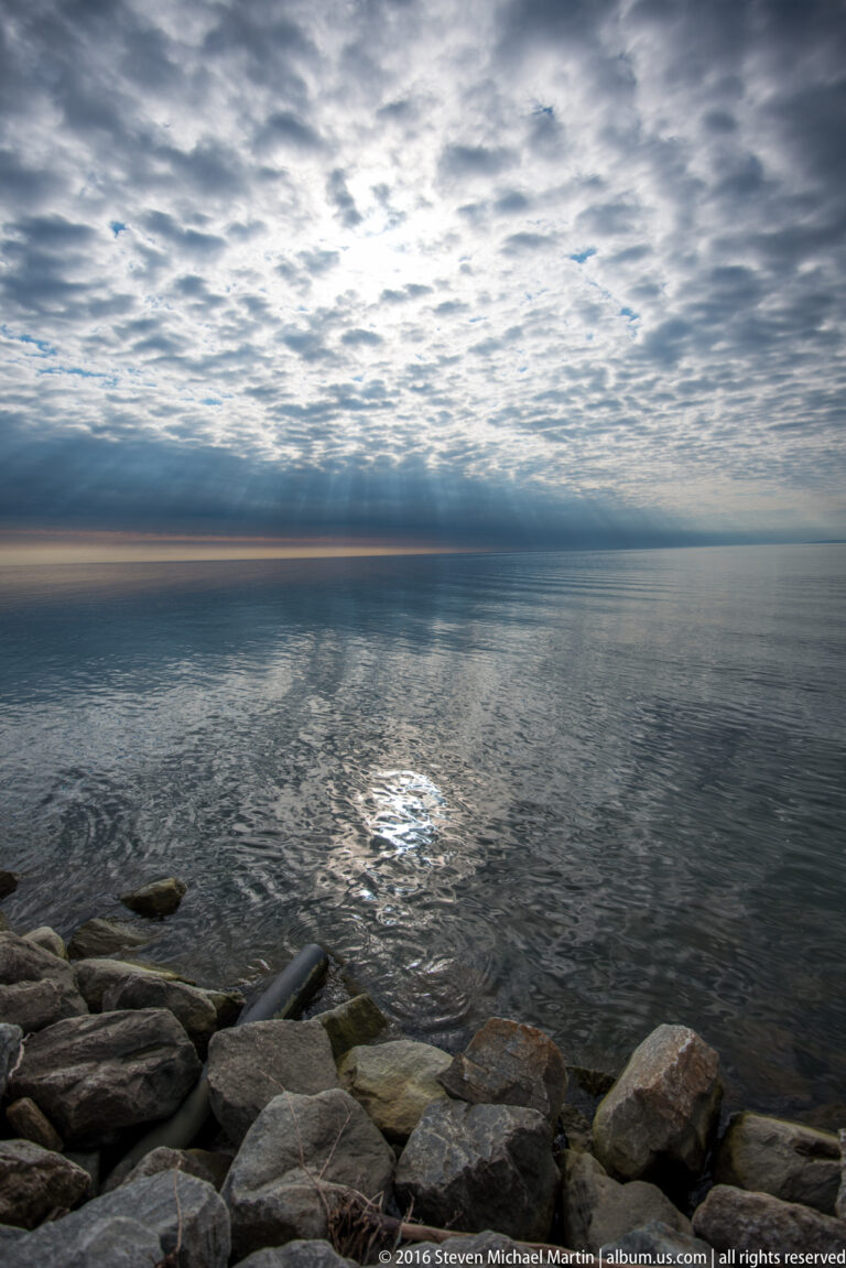 A bright reflection shimmers on rippled water beyond rounded shoreline stones beneath a broken cloud cover.