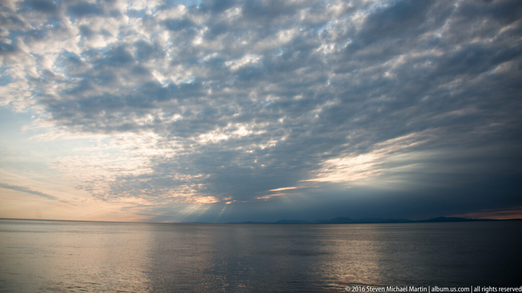 Sunlight breaks through rippled clouds above open water, reflecting near a low dark coastline.