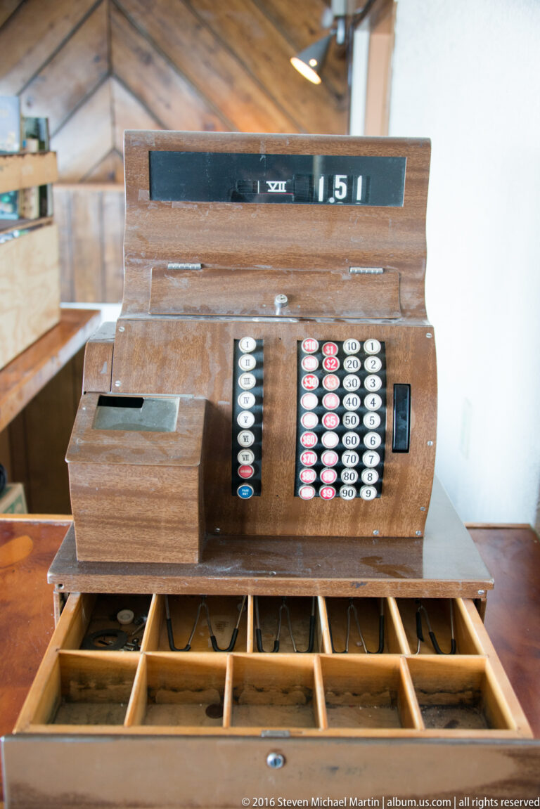 A wooden mechanical cash register with rows of keys sits above an open, divided cash drawer.