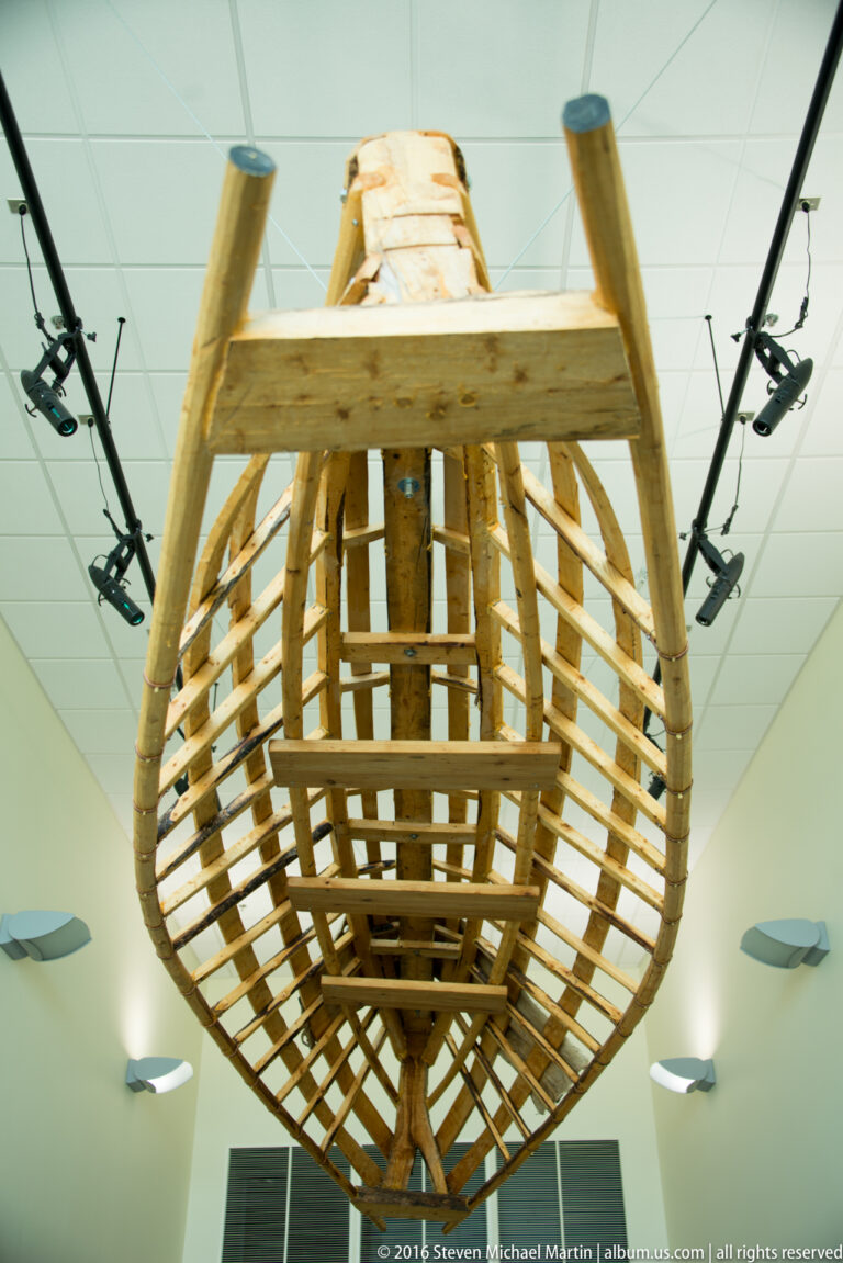 A narrow wooden boat frame stands upright, exposing its curved ribs and lengthwise supports.