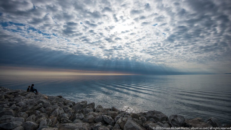 Two people sit on a rocky breakwater beneath a wide clouded sky with sunbeams over calm water.