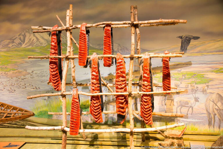 Split fish hang in rows from a wooden drying rack in front of a painted landscape.