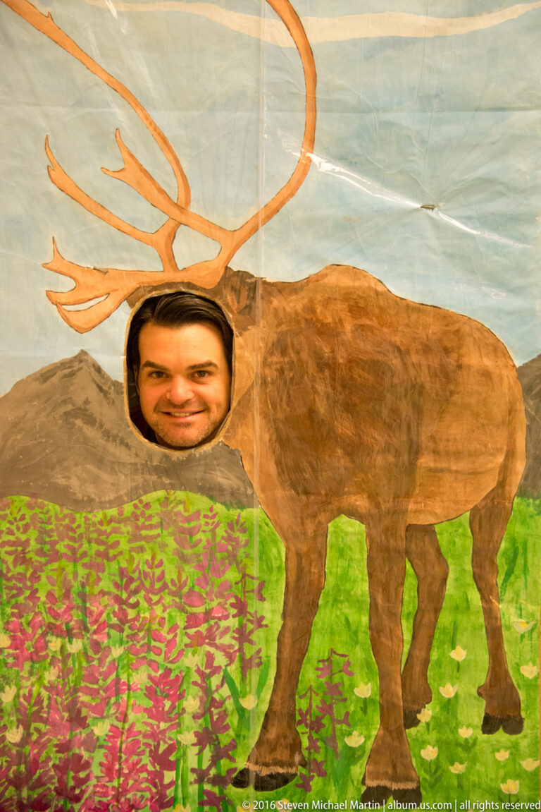 A visitor looks through a face opening in a painted moose panel with large antlers.