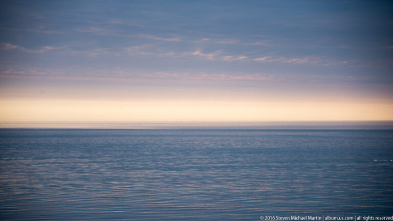 A narrow pastel sunset glows above a level horizon, with fine ripples across the blue water.