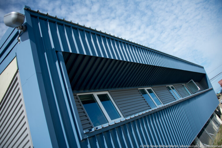 A blue-gray metal-clad building rises diagonally beneath a cloudy sky, with tall windows along the facade.