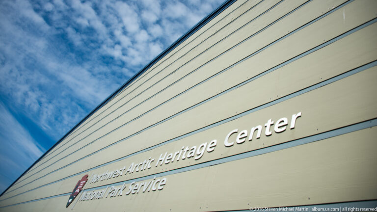 The Northwest Arctic Heritage Center name and National Park Service emblem appear on pale siding beneath a blue sky.