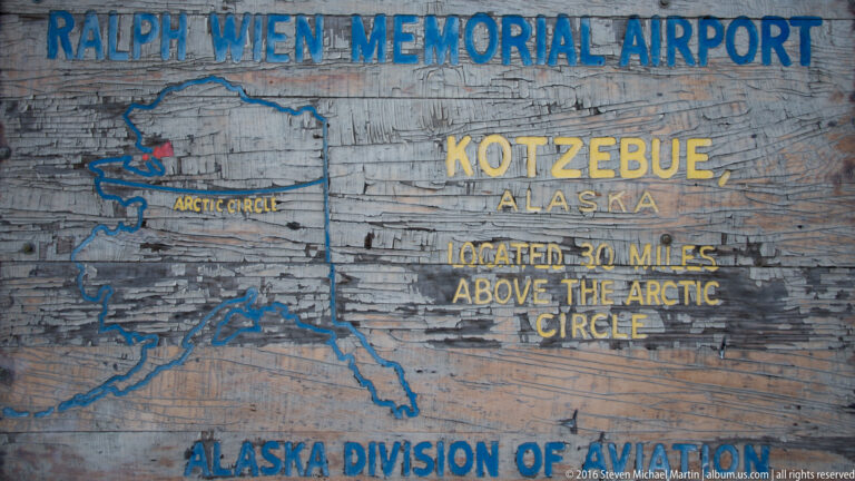 A weathered wooden sign names Kotzebue and the Wiley Post–Will Rogers Memorial Airport, with a map and Arctic Circle note.