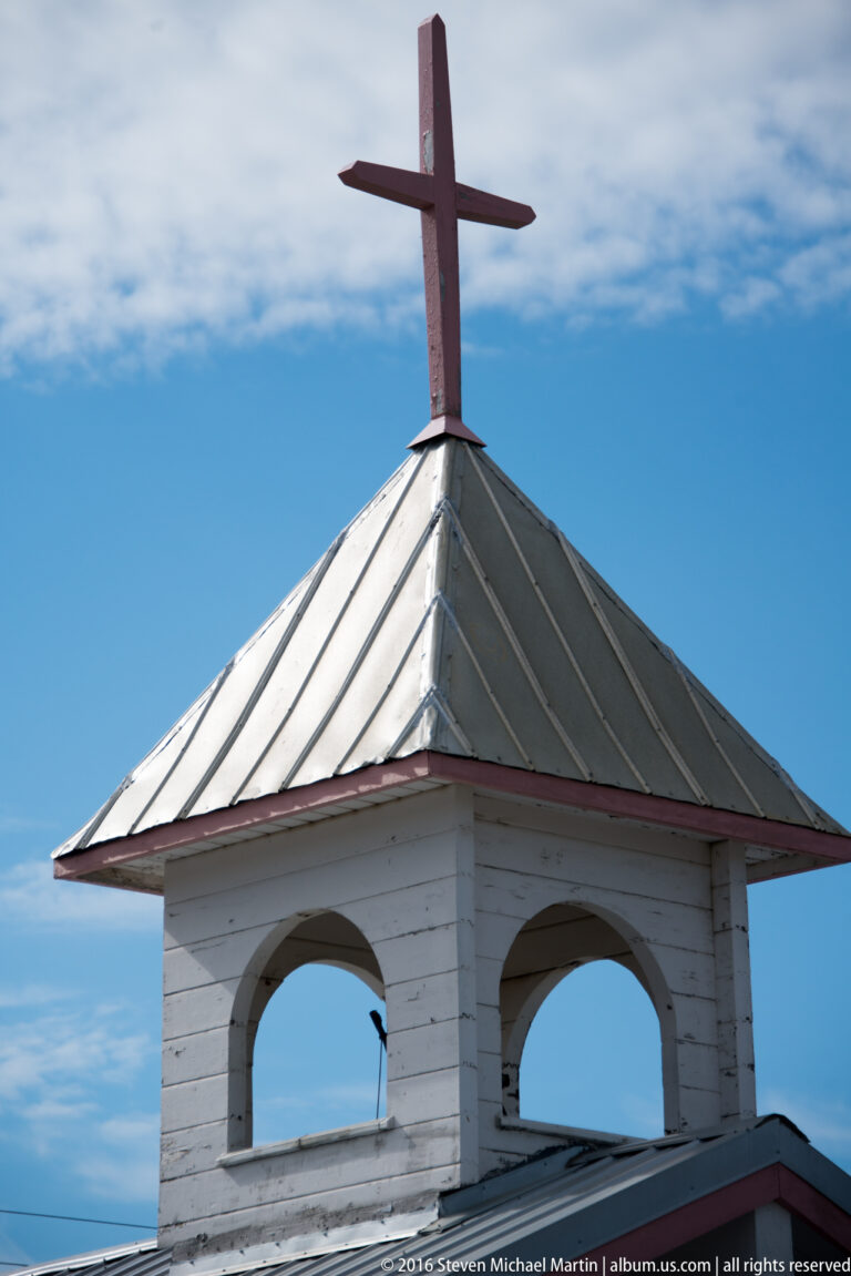 A tall cross stands above the metal roof of a small white open-sided structure.