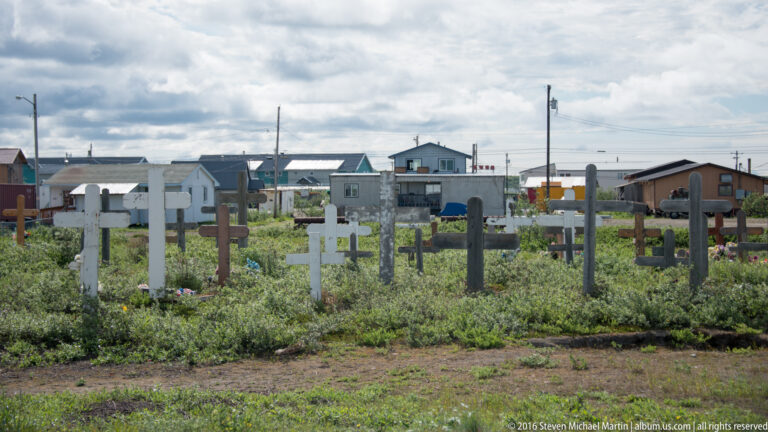 Rows of crosses and grave markers rise from a grassy cemetery with houses behind them.