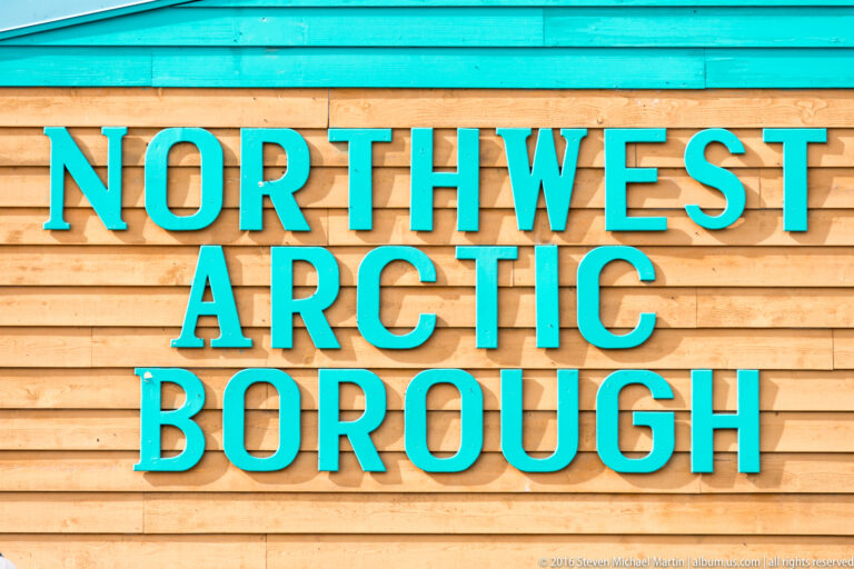 Turquoise letters spelling “Northwest Arctic Borough” are mounted on horizontal wooden siding.