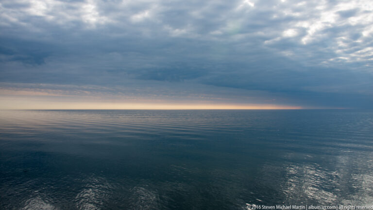 Dark clouds hang over smooth open water, with a small bright reflection below a narrow sunset band.