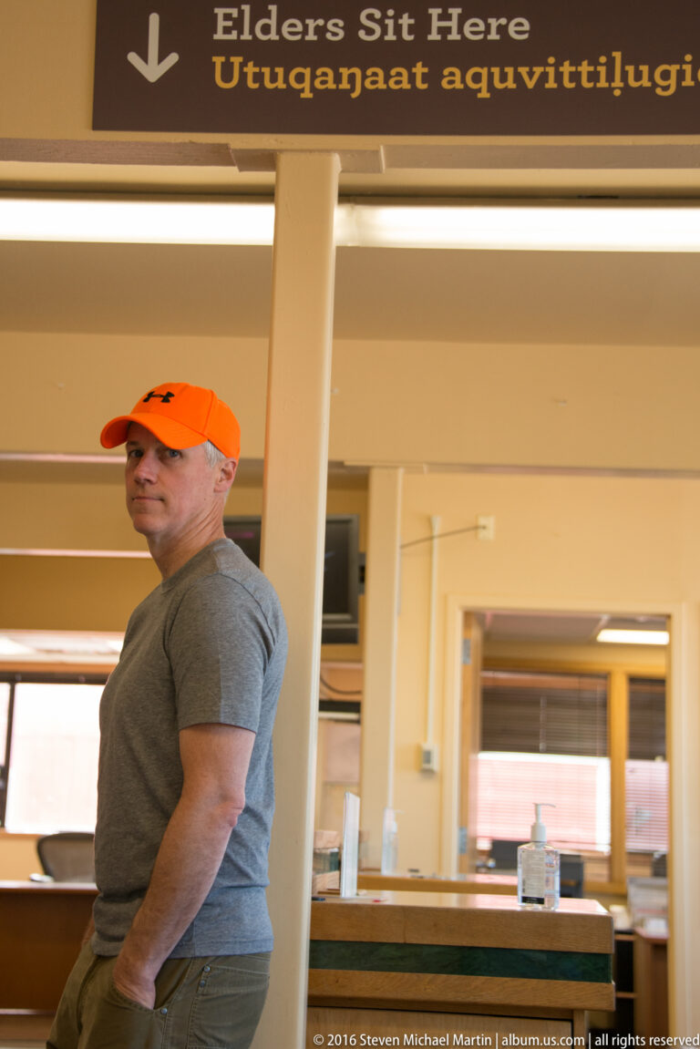 A man in an orange cap stands beneath an “Elders Sit Here” sign with Iñupiaq text in a bright interior.