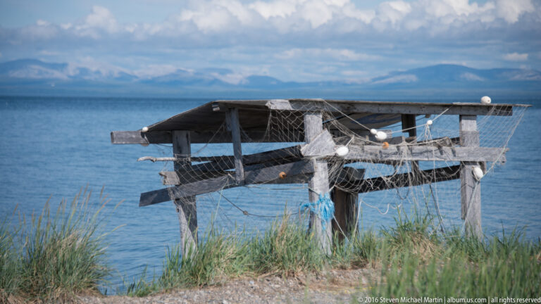 Netting and pale floats hang from a wooden rack beside calm water and grassy shore.