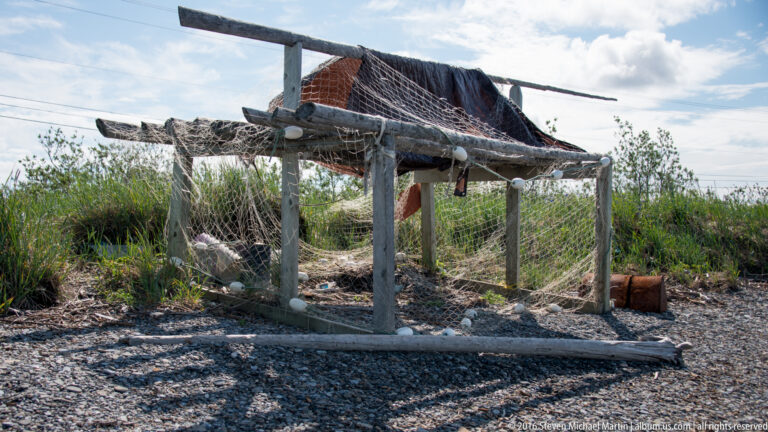 A raised timber frame draped with netting stands on a gravel bank beside shrubs and low buildings.