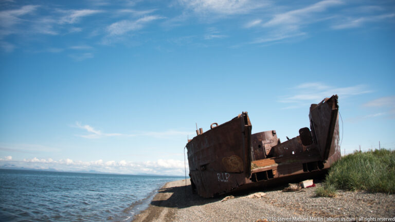 A rusted steel vessel with an open damaged hull sits at an angle on a narrow beach.