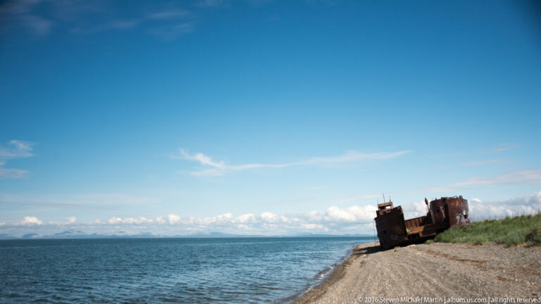 A rusted vessel rests on a narrow coastal spit between open water and a grassy bank.
