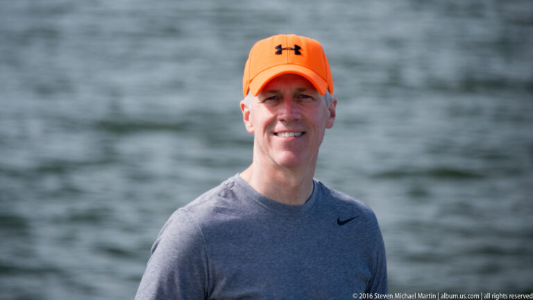 A smiling man in a bright orange cap and gray shirt poses before softly blurred water.