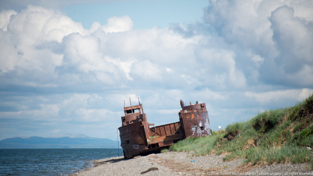 A rusted vessel sits on a narrow beach beneath large clouds, between open water and a grassy bank.