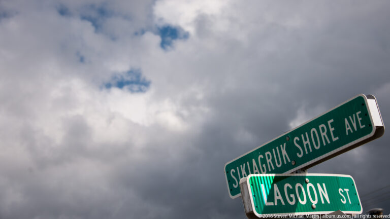 Green street signs for Shagluk Shore Avenue and Lagoon Street stand against a cloudy sky.