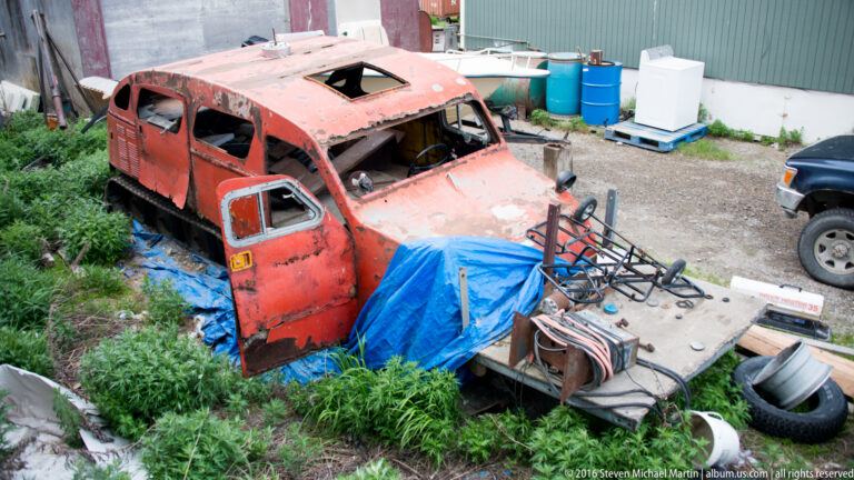 A rusted red vehicle cab sits among tires, metal parts, a blue tarp, and barrels in a yard.