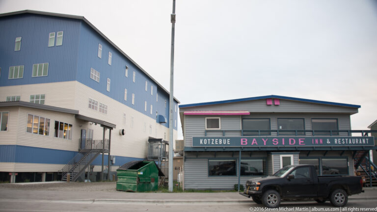 A pickup and dumpster stand beside the Kotzebue Bayside Inn and Restaurant and a taller blue-and-white building.
