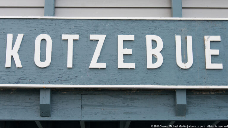 Raised white letters spelling Kotzebue cross a weathered blue signboard.