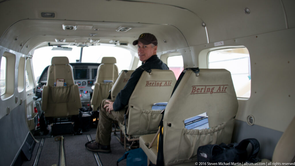 A man sits inside a small Bering Air plane behind pale passenger seats, with the cockpit visible ahead.