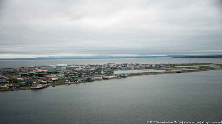 An aerial view shows Kotzebue’s buildings and roads along a narrow spit between lagoon and open water.
