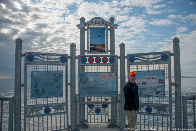A visitor stands beside a three-part metal gateway labeled Kotzebue, with the sea visible behind it.