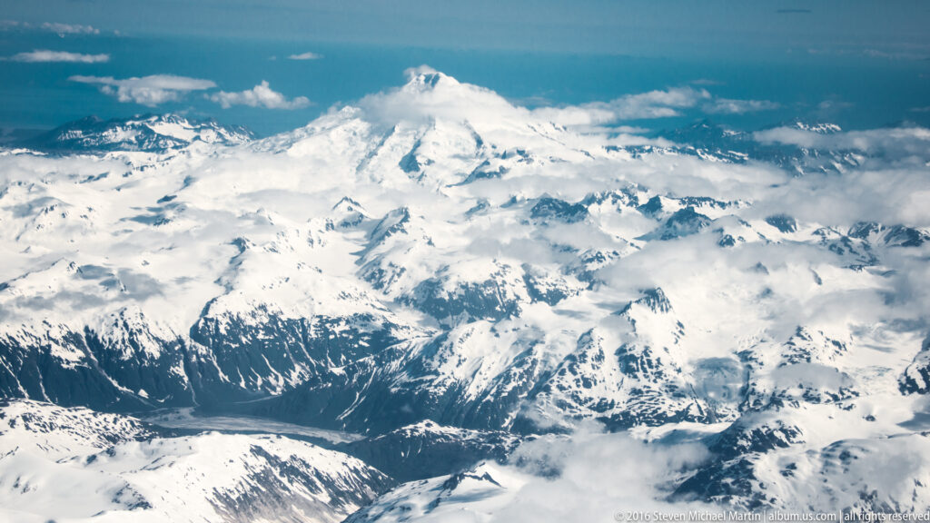 A snow-covered mountain towers above a dark valley and surrounding peaks, with clouds crossing the lower slopes.