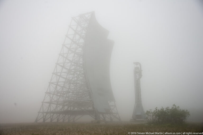 Two White Alice antenna structures appear as faint silhouettes in dense fog above low vegetation.