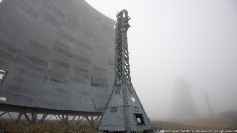 A riveted steel mast stands beside a curved White Alice reflector as another structure fades into fog.