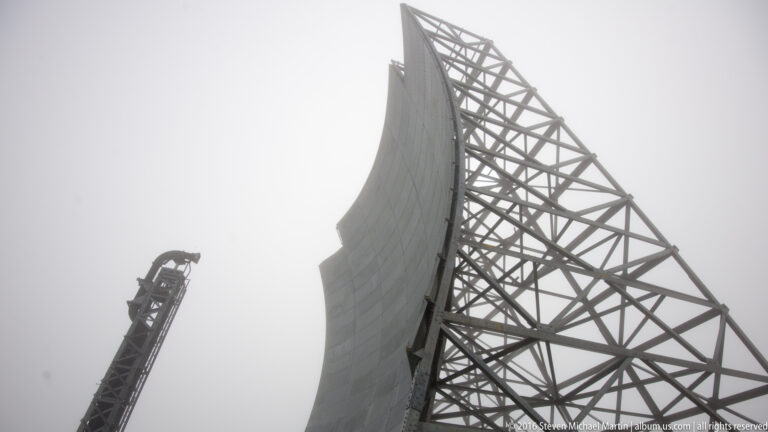 A steel lattice tower rises beside the curved edge of a White Alice reflector against fog.