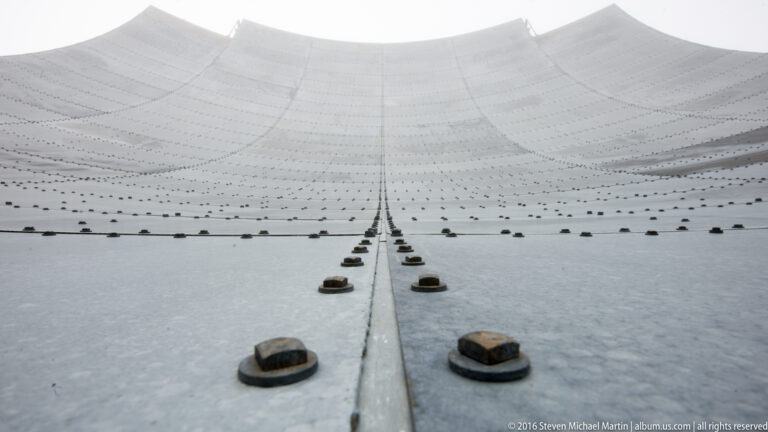 Rows of fasteners and a raised seam cross the curved metal face of a White Alice reflector.