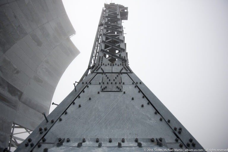 Rows of bolts climb a triangular lattice mast framed by cropped White Alice reflector panels.