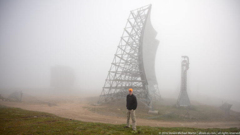 A visitor in an orange cap stands below White Alice antenna structures on a foggy slope.