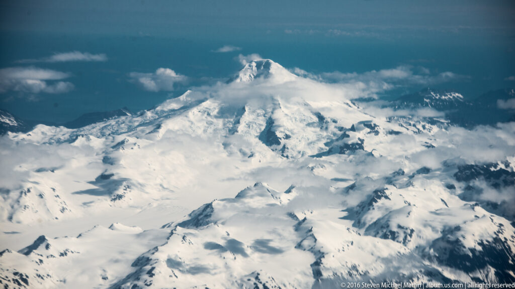 A pointed snow-covered summit rises above glaciers and dark rocky ridges beneath scattered clouds.
