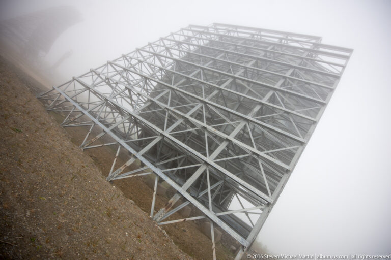 A White Alice antenna framework angles across a gravel ridge on triangular steel legs.