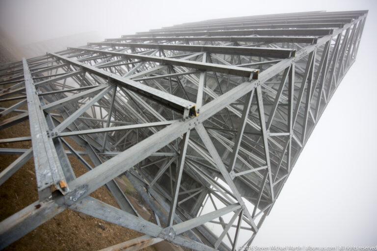 Intersecting beams and parallel trusses extend beneath a White Alice reflector above a gravel slope.
