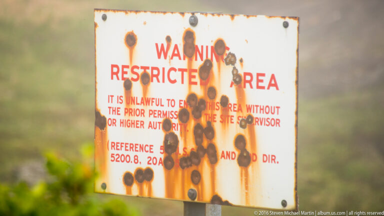 A rust-streaked warning sign reads “Restricted Area,” with dark marks obscuring smaller text.