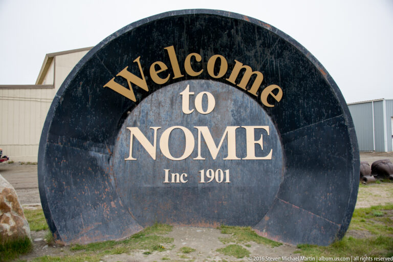 A curved dark sign on grass reads “Welcome to Nome Inc. 1901” in large gold and cream letters.