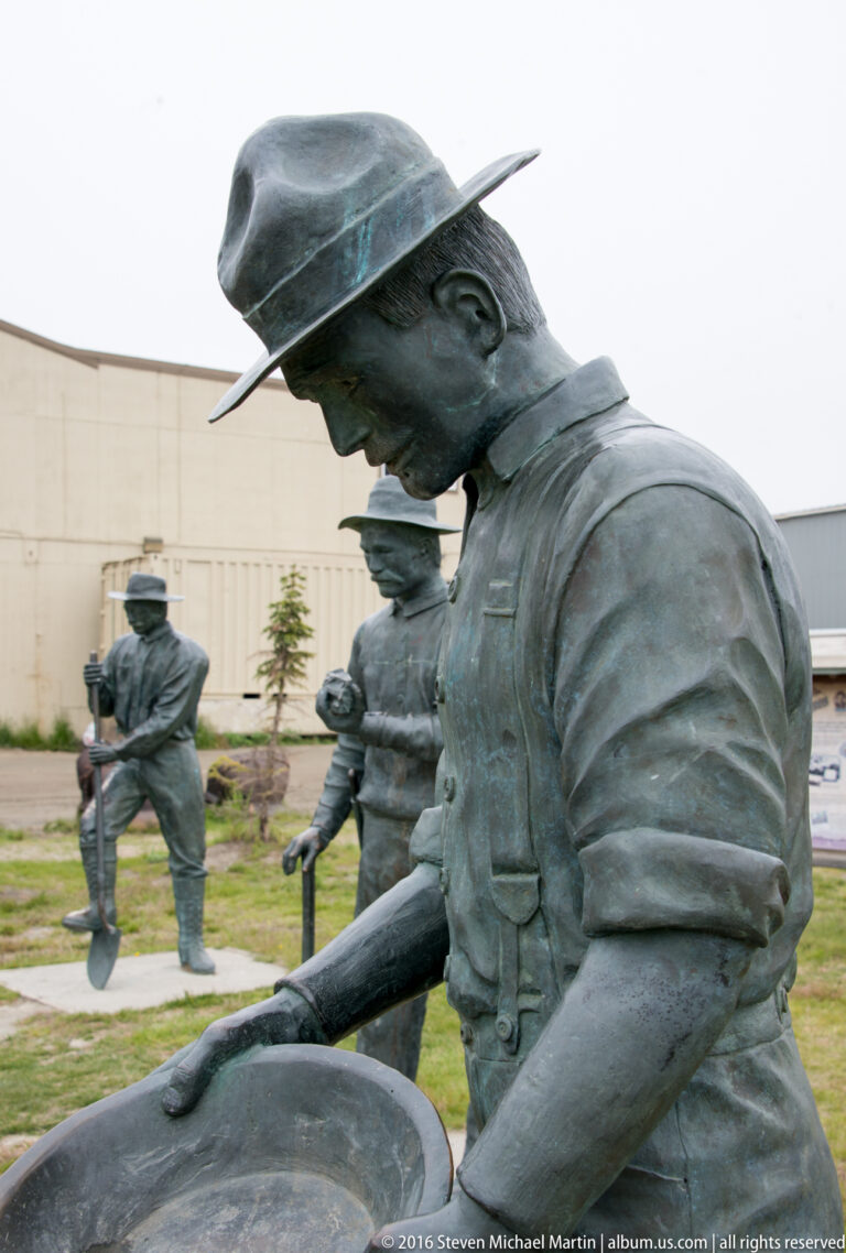 A bronze prospector bends over a pan, with other figures and mining tools behind him.
