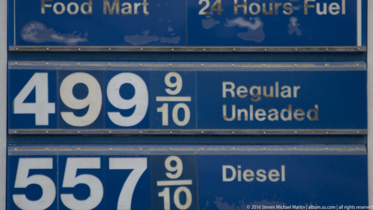A blue price board lists regular unleaded at 499 9/10 and diesel at 557 9/10 beneath a 24-hour fuel header.
